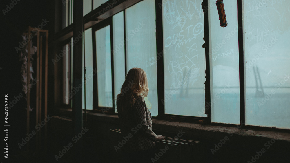 Wall mural The young woman watching out of the window in an abandoned factory located in Saint Petersburg, Russia.