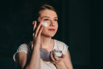 Happy young woman doing beauty treatment. The girl applies white cream on her face on the morning.