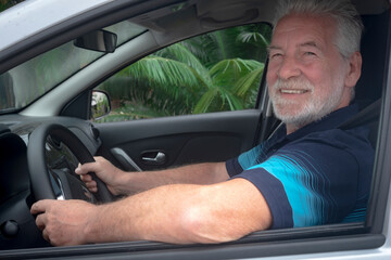 Attractive senior man smiles after parking the car. Caucasian senior people with gray hair and beard. Green plants outside the car window
