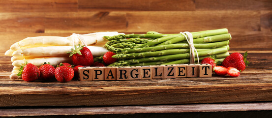 White and green asparagus and strawberries on background with the german word Spargelzeit in wooden letter. - 354728234