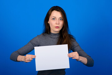 Young woman in grey shirt holding a sheet of paper and looking at the camera, smiling. Blue background isolated