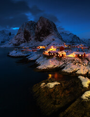 Famous tourist attraction Hamnoy fishing village on Lofoten Islands, Norway with red rorbu houses in winter snow illuminated in the evening.
