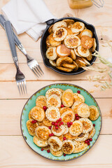 Trend breakfast. Dutch mini pancakes with red currants on a plate and a pan with them on a wooden table. Vertical. Top view
