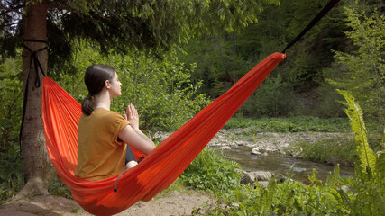 Happy serene woman meditates in a hammock sitting in namaste pose outdoor. Concept of meditation, relaxation, spirituality and recreation at the nature. 