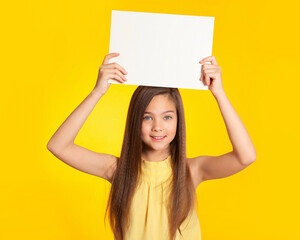 Beautiful European brunette teenager girl with blue eyes holds a white sheet of paper over her head on a yellow background and smiles. Place for text, banner, surface for inscriptions and advertising.