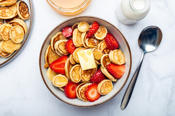 Trendy home breakfast with tiny cereal pancakes with piece of butter and fresh strawberries in the bowl. View from above.