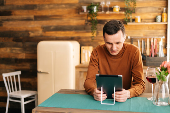 Young cheerful man using tablet computer having online video chat with friends while sitting at the table in cozy room.