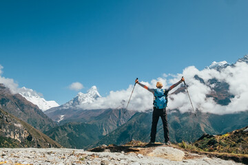 Active vacations concept image.Young hiker backpacker man rising arms with trekking poles enjoying the Thamserku 6608m mountain during high altitude Acclimatization walk. Everest Base Camp route,Nepal