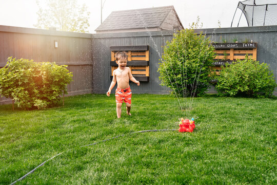 Kid's outdoor activity. Funny toddler boy wearing a orange swimming shorts running and jumping around garden sprinkler playing with water splashes having fun in the backyard on a sunny hot summer day.