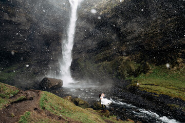 The groom circles the bride in his arms near the waterfall, it is snowing. Destination Iceland wedding, near Kvernufoss waterfall.
