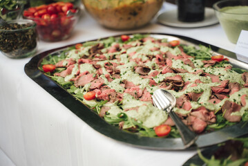 Stainless tray with slices of roast beef poured with green cream sauce. Self-service buffet table. Celebration, party, birthday or wedding concept.