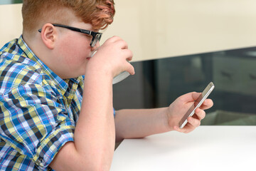 Red-haired boy with cellphone drinking from a glass of ice water with lemon. Child with glasses dressed in a plaid shirt looks at the smartphone and drinks water.