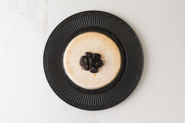 Top view of a coconut pudding with plums in a black plate in a marble table and space for text