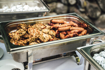Stainless hotel pan with spiced chicken steaks and grilled sausages. Self-service buffet table. Celebration, party, birthday or wedding concept.