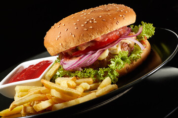 Big fresh burger with sauce and fries on black plate isolated on black background.  American classic burger menu.