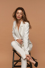 Girl in white suit with loose dry hair and make-up sitting on high chair and posing on a beige background. Fashion style body length studio portrait. Elegant fashionable woman