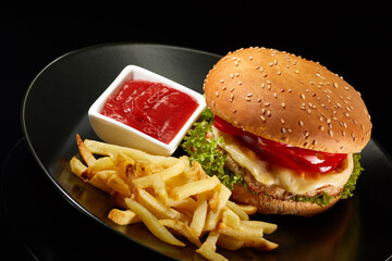 Big fresh burger with sauce and fries on black plate isolated on black background.  American classic burger menu.