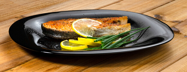 Grilled salmon steak with lemon slice and herbs in a black plate on a wooden background