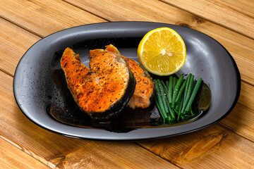 Grilled salmon steak with lemon slice and herbs in a black plate on a wooden background