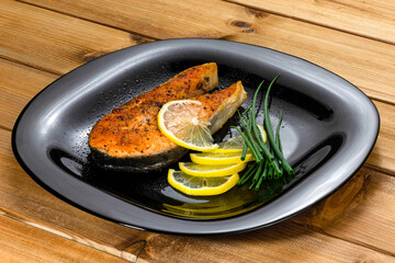 Grilled salmon steak with lemon slice and herbs in a black plate on a wooden background