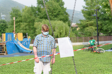 Boy wearing surgical mask goggles dressed in a plaid shirt in front of a red and white striped barrier tape around the playground.Child in front of a red and white stripe warning tape in a public park