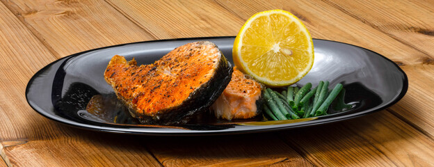 Grilled salmon steak with lemon slice and herbs in a black plate on a wooden background
