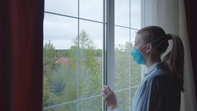 Girl in pajamas and a blue protective mask goes to the window and looks sadly at the street, sighs heavily. Girl in insulation complies quarantine
