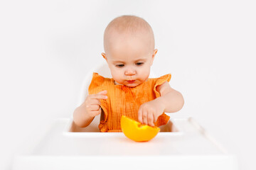 Happy baby girl sitting in high chair eating oranges in a white kitchen. Healthy nutrition for kids. Bio fruits as first solid food for infant.