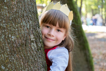 Portrait of a pretty smiling child girl in a paper crown on her head leaning to a tree in summer...