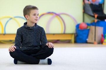 Young child boy sitting and relaxiong on the floor inside sports room in a school after training.