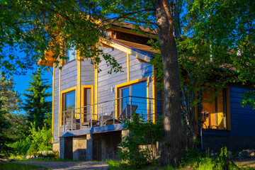Wooden cottage with a terrace. House among the trees. Stay in the fresh air. Outdoor veranda with sun lounger, chairs and table. Rent cottages. Rest in Karelia. Sunny summer day outside the city.