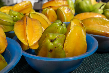 Starfruit both yellow and green sitting in small blue plastic bowls on a table at a farmer's market. Six pieces of fruit in each bowl. The skins are waxy, shiny and thick. 