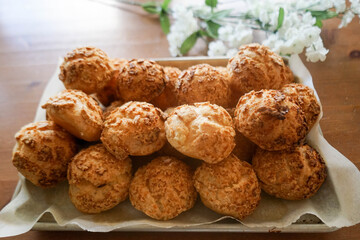 Homemade Choux Au Craquelin (crispy cookie shell-covered cream puffs) stacked in rows on a baking tray and a well sun-lit table. White flowers in bokeh at the back for decoration.