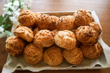 Homemade Choux Au Craquelin (crispy cookie shell-covered cream puffs) stacked in rows on a baking tray and a well sun-lit table. White flowers in bokeh at the back for decoration.