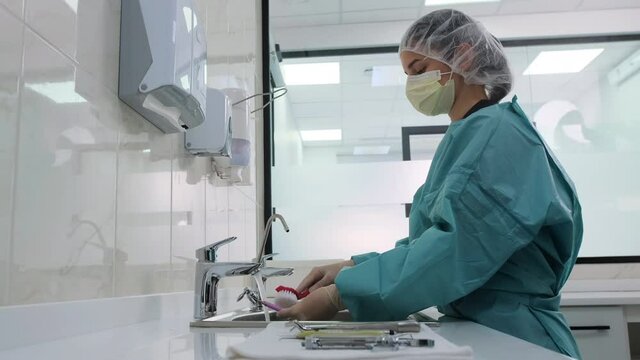 Side view of woman laboratory technician in white gloves wash dental medical instruments with a red brush under a stream of running water. Processing, disinfection of dental probe.