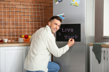Young man writing text I LOVE YOU on chalkboard in kitchen