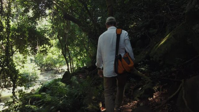 A man in a white shirt walks next to a river through a jungle