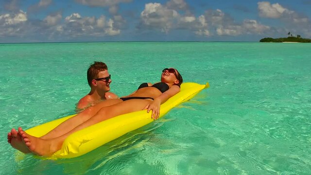 Zoom-in View Of A Caucasian Lady Lying On An Inflatable Yellow Raft, Assisted By Her Boyfriend On A Beautiful Tropical Island.