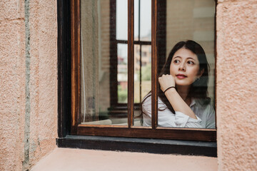 view through window glass young beautiful asian korean woman tourist sitting in old house and looking out with beautiful sunny day. close up lady traveler with cute charming attractive face indoors.