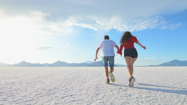 Bonneville Salt Flats near Salt Lake City, Utah with back view slow motion of young man woman happy couple running jumping on white field in summer holding hands