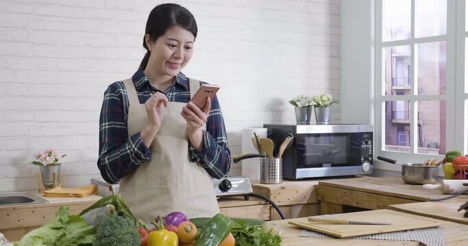slow action of young beautiful wife in apron holding cellphone text message to husband asking if having dinner at home. japanese woman smiling using mobile phone have conversation in wood kitchen