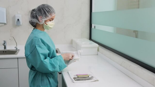 Woman doctor assistant in white gloves and green gown is wiping odontic instruments after washing, before sterilization and puts on the tablecloth in the dental office.