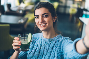 Cheerful young woman holding tasty cocktail in hand and making selfie photo on modern smartphone device
