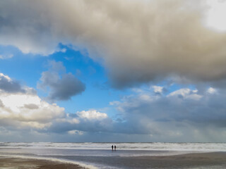 Couple of people standing on sandy beach shoreline with many waves on a day with big clouds