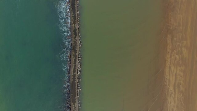 A top-down view of the  Cantabrian Sea on the coast of Spain shows a contrasting line of a breakwater separating the darker tones of the deep water from the earthy browns of the land and sand.