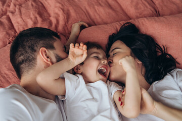 Close up. Pregnant mother playing with her child in the bedroom.Family are sharing good emotions while relaxing at home Big window, natural light, terracotta bed linen. Have fun, kiss and hugs.