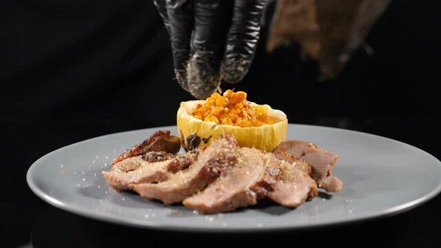 Serving food concept. Chef maiking final decoration of dish with sliced meat and baked pumkin with stuffing. Cook sprinkles meat with salt, pepper, seasoning, spices and brown sugar. Slow motion. Full