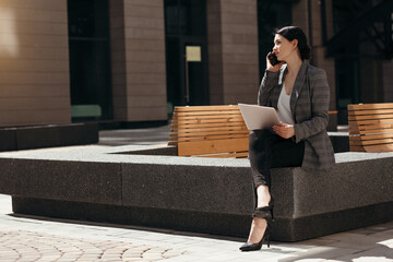 Rear view of a business woman in a jacket sitting on a bench with a laptop on her lap, working...