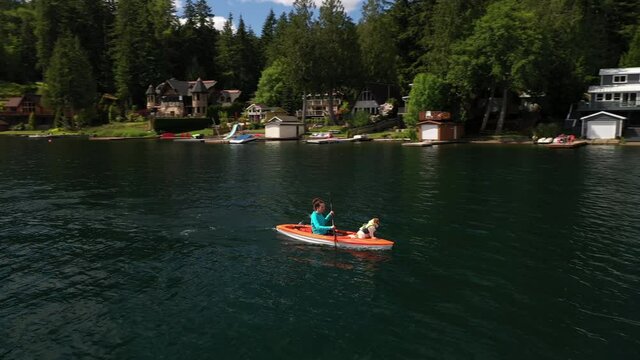 Young woman and her dog rowing a canoe on the water with a view of lake houses in the distance; 4k 24fps clip