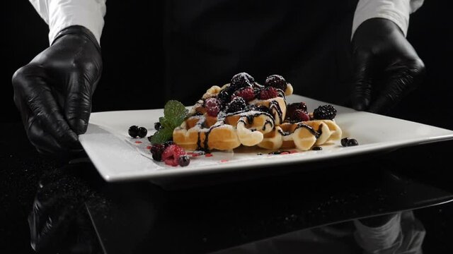 Restaurant food concept. Chef serves prepared tasty dessert. Delicious waffles with berries and chocolate topping on black background. Male cook presenting dish. Slow motion. Full hd